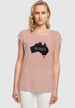 Rosa t-skjorte med korte ermer som har en svart kontur av Australia med ordet "AUSTRALIA" i fet skrift. Myk tekstur av stoffet.