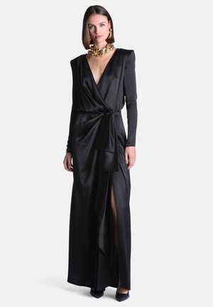 Femme portant une longue robe portefeuille en satin noire avec un décolleté en V profond, manches longues, fente sur la cuisse, et un collier et des boucles d’oreilles dorés et épais, debout sur fond blanc.