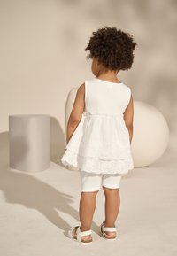 White sleeveless dress with lace trim over white ribbed shorts, paired with white sandals. Smooth fabric with layered design.