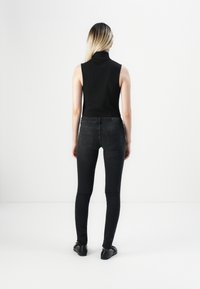 Žena s dlouhými blond vlasy, oblečená v černém bezrukávovém roláku a černých skinny džínách, stojící čelem k bílé zdi.