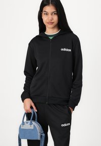 Fekete zipzáros kapucnis pulóver fehér Adidas logóval, fekete melegítőnadrággal párosítva. Egy kék táska van a kezében, amelynek dupla fogantyúja és vállpántja van.