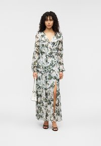 Μάξι φόρεμα από διαφανές ύφασμα με λουλουδάτο μοτίβο σε πράσινο και άσπρο, με ντεκολτέ σε σχήμα V, μακριά μανίκια και σκίσιμο μέχρι το μηρό, συνδυασμένο με άσπρη τσάντα.