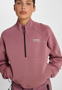Lyserød quarter-zip sweatshirt med høj krave, der har en sort lynlås og "hummel"-logoet printet i hvidt på brystet.
