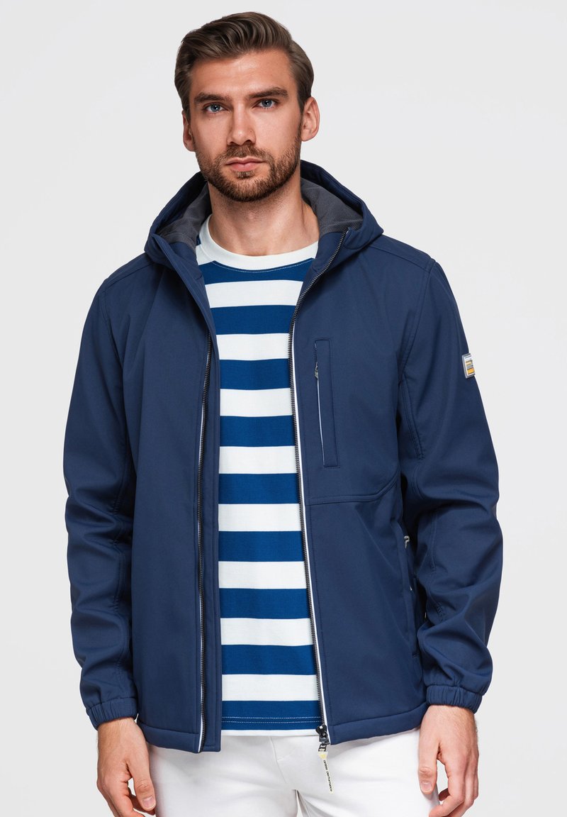 Homme portant une veste à capuche bleue sur une chemise blanche rayée bleue et un pantalon blanc, debout devant un fond uni.