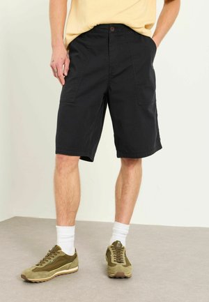 Mann trägt schwarze knielange Shorts mit Fronttaschen, ein gestecktes gelbes T-Shirt, weiße Socken und olivgrüne Sneakers und steht auf einem grauen Boden.