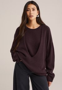 Üks suur suuruses, sügava V-kaelusega kootud sviiter rikkaliku burgundi värviga, millel on pikad varrukad ja vabalt langev lõige, koos tumedate pükstega.