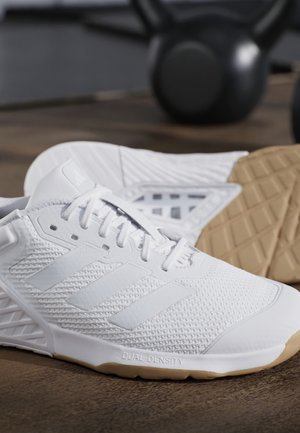 Biały sportowy but Adidas z gumową podeszwą i cholewką z siateczki, umieszczony na drewnianej podłodze obok czarnych odważników kettlebell.