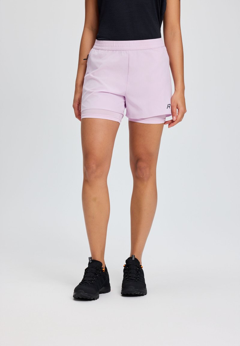 Mujer con pantalones deportivos deportivos morados claros en capas y zapatos de senderismo negros, de pie contra un fondo liso gris claro.