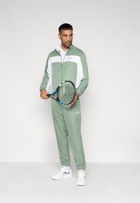 Σετ φόρμας σε ανοιχτό πράσινο και λευκό, με μπουφάν με φερμουάρ και ελαστικά τελειώματα, ταιριαστό παντελόνι και ρακέτα τένις. Περιλαμβάνονται λευκά αθλητικά παπούτσια.