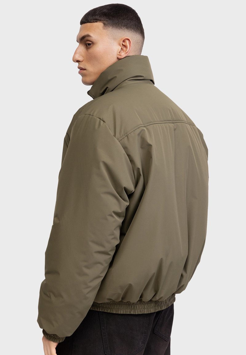 Giacca bomber imbottita in verde oliva. Presenta un colletto alto, polsini elasticizzati e orlo con cordino. Texture liscia con dettagli di cucitura minimi.