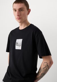 T-shirt in cotone nero con maniche corte e scollo rotondo, caratterizzato da una stampa grafica testurizzata sul petto e un'etichetta rossa.