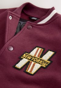 Bordeauxrotes Collegejacke mit einem strukturierten, gelb-weißen gestickten Logo mit der Aufschrift "NEW YORK CITY" und Streifen, silbernen Druckknöpfen und gestreiftem Kragen.