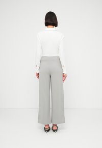 Γυναίκα που στέκεται με την πλάτη προς την κάμερα, φοράει λευκό μπλουζάκι μακρυμάνικο, γκρι φαρδύ παντελόνι και μαύρα τακούνια με ανοιχτή πλάτη, μπροστά από ένα απλό φόντο.