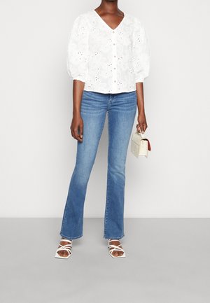 Bílá květinová krajková halenka s výstřihem do V, nafouklými rukávy a knoflíkovým detailem, v kombinaci s rozšířenými modrými džínami a bílými sandály na tenkých remíncích.