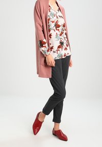 Light pink long cardigan over a floral white top, dark gray pants, and red pointed shoes. Textured cardigan with loose fit and floral pattern.