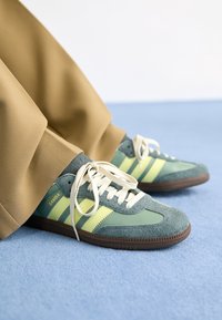 Green suede and leather sneakers with yellow stripes, flat laces, and a brown rubber sole, worn with beige pants on a blue surface.