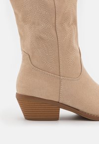 Beige suède cowboy-ankelboot met decoratieve stiksels en een gevormde middenhak met een ribbelstructuur.