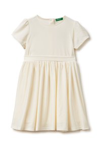 Kremowa sukienka dla dziewczynki z krótkim rękawem, z marszczonym pasem i spódnicą, okrągłym dekoltem oraz małą zieloną metką marki wewnątrz kołnierzyka.