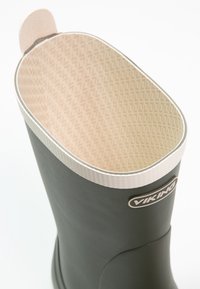 Botas de goma verdes con un interior texturizado de color beige claro, que cuentan con un borde blanco y una etiqueta de logo. Superficie exterior lisa y parte superior redondeada.