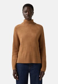 Vrouw in een camelgekleurde coltrui met een geruit patroon en ribbels aan de mouwen, gecombineerd met donkerblauwe denimjeans, staand in een neutrale houding.