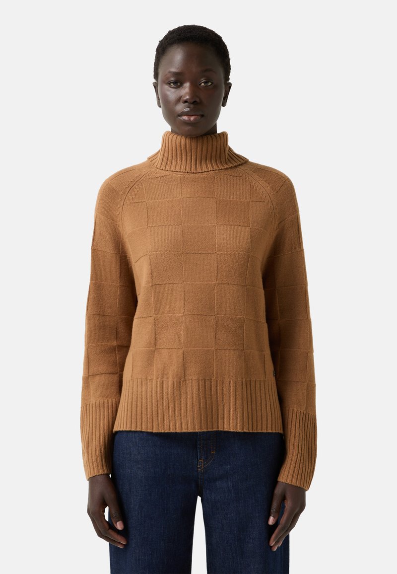 Vrouw in een camelgekleurde coltrui met een geruit patroon en ribbels aan de mouwen, gecombineerd met donkerblauwe denimjeans, staand in een neutrale houding.
