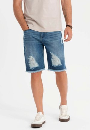 Jeanshorts i mellemblå med slidte detaljer og ophavede kanter, parret med hvide sko. Glat stofstruktur, afslappet design.