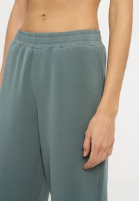 Γυναίκα που φοράει φαρδιά γκρι-πράσινα φούτερ παντελόνια με ψηλή μέση και ελαστική μέση, κομμένα στο ύψος της κοιλιάς, με το δεξί χέρι ακουμπισμένο στο πλάι.