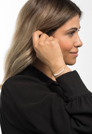 Goldenen Armband mit kleinen weißen Perlen, das am Handgelenk getragen wird. Der Hintergrund ist schlicht in weiß gehalten, während eine dunkle Bluse teilweise sichtbar ist.