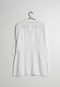 Camicia bianca in lino a maniche lunghe appesa a un gruccia, con scollo rotondo, vestibilità rilassata e una texture leggermente trasparente. Fessure laterali all'orlo.