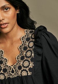Blusa nera con intricati motivi floreali ricamati in beige, scollatura a onda e maniche a sbuffo realizzate in tessuto testurizzato.