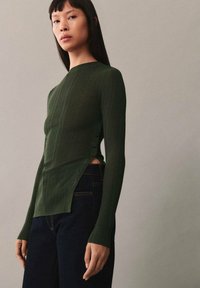 Dunkelgrünes Langarmshirt mit geripptem Muster, hohem Kragen und seitlichen Knopfakzenten, kombiniert mit dunklen Jeans.