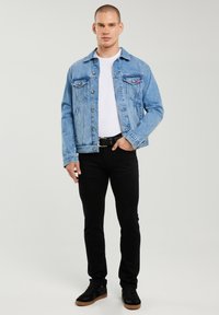 Mann steht und trägt eine hellblaue Jeansjacke, weißes T-Shirt, schwarze Hose und schwarze Sneaker mit gumfarbener Sohle vor grauem Hintergrund.