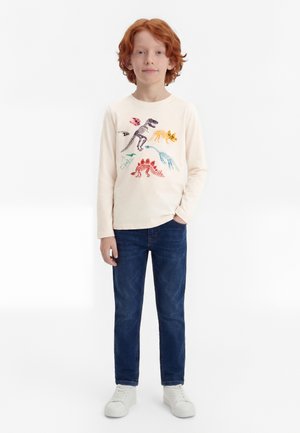 Niño con cabello rojo que lleva una camiseta de manga larga color crema con estampados de esqueletos de dinosaurios de colores, jeans azules y zapatillas blancas, de pie con una mano en el bolsillo.