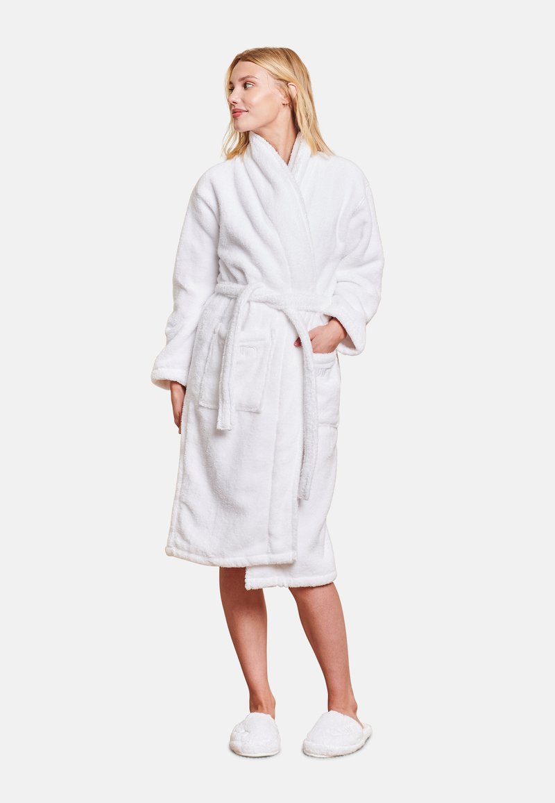 Witte, zachte badjas van badstof met een sjawlboord, een ceintuur in de taille en twee voorzakken. Het model draagt pluizige witte slippers.