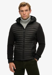 Mann in schwarzer, gesteppter Kapuzenjacke über einem grauen Rollkragenpullover, der mit einer Hand in der Tasche vor weißem Hintergrund steht.