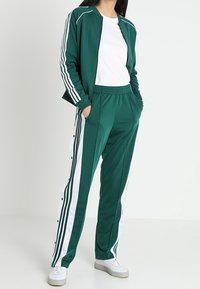 Zelená tepláková souprava s bílými postranními pruhy, kalhoty s knoflíkovými detaily a lehká bunda na zip přes bílé tričko.