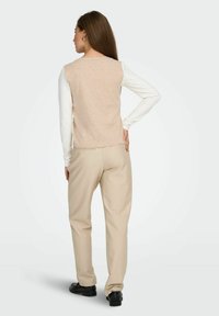 Top senza maniche in maglia beige indossato sopra una camicia bianca a maniche lunghe, abbinato a pantaloni beige e scarpe nere. Texture liscia, design semplice.