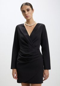 Robe noire portefeuille à manches longues, en tissu texturé, décolleté en V profond et détail froncé à la taille ; accompagnée d'un collier en perles.