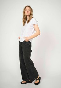 Vit kortärmad t-shirt kombinerad med hög midja svarta vida jeans. Modellen står avslappnat med händerna i fickorna och har på sig svarta sandaler.