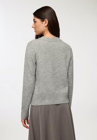 Donna con lungo capelli scuri, che indossa un maglione grigio chiaro e una gonna beige ampia, vista da dietro su uno sfondo neutro.