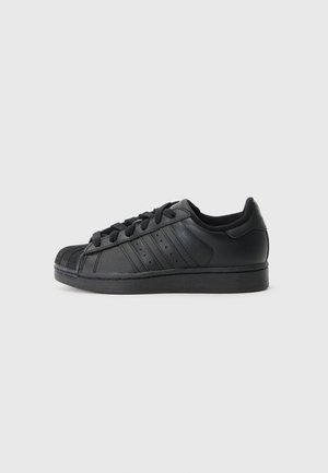 Sort lavtop sneaker med gummiskjoldetå, snørelukning, tre sideløb og flad sål mod en grå baggrund.