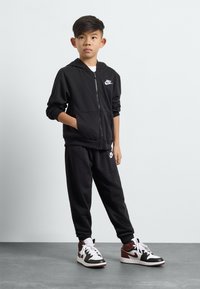 Črna zip-up kapuca in ustrezne trenirke, obe z belimi logotipi Nike. Temni športni copati z črnimi, belimi in maroon akcenti. Gladka tekstura tkanine.