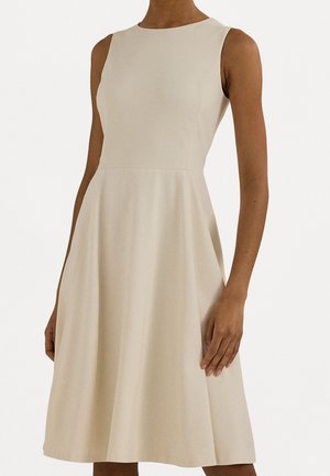 Robe beige sans manches, longueur au genou, avec corsage ajusté et jupe évasée, portée par une personne debout sur fond blanc.