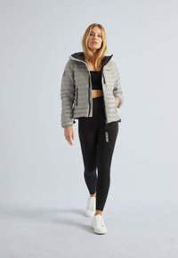 Grå pufferjakke med lynlås foran, letvægtsdesign og kontrastsort foring. Bæret med sorte leggings og hvide sneakers.