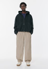 Fekete, cipzározható kapucnis pulóver bézs kordbársony nadrággal és krémszínű sportcipőkkel párosítva. A modell fehér háttér előtt áll.