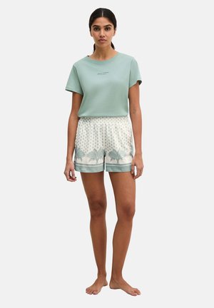 Femme debout pieds nus portant une chemise à manches courtes vert clair et un short blanc avec des motifs de palmiers verts.