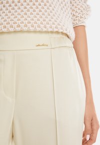 Light beige tailored pants with a smooth texture, elastic waistband, and a gold logo accent on the front, featuring sharp pleats.