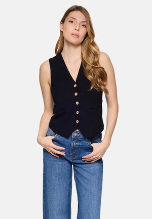 Mujer con cabello largo y ondulado, vestida con un chaleco negro abotonado y vaqueros azules, de pie con las manos en los bolsillos contra un fondo liso.