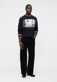 Homme debout, main dans la poche, portant un sweat-shirt noir avec des rayures blanches et du texte, un pantalon noir et des chaussures noires sur un fond blanc.