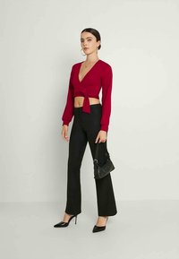 Mulher vestindo uma blusa vermelha de mangas longas e cropped, calças pretas de cintura alta, saltos pretos e segurando uma pequena bolsa preta.